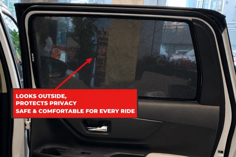 A fitted rear-door window shade seen from inside the car. The street, parked cars and shopfronts outside are clearly visible through the mesh.